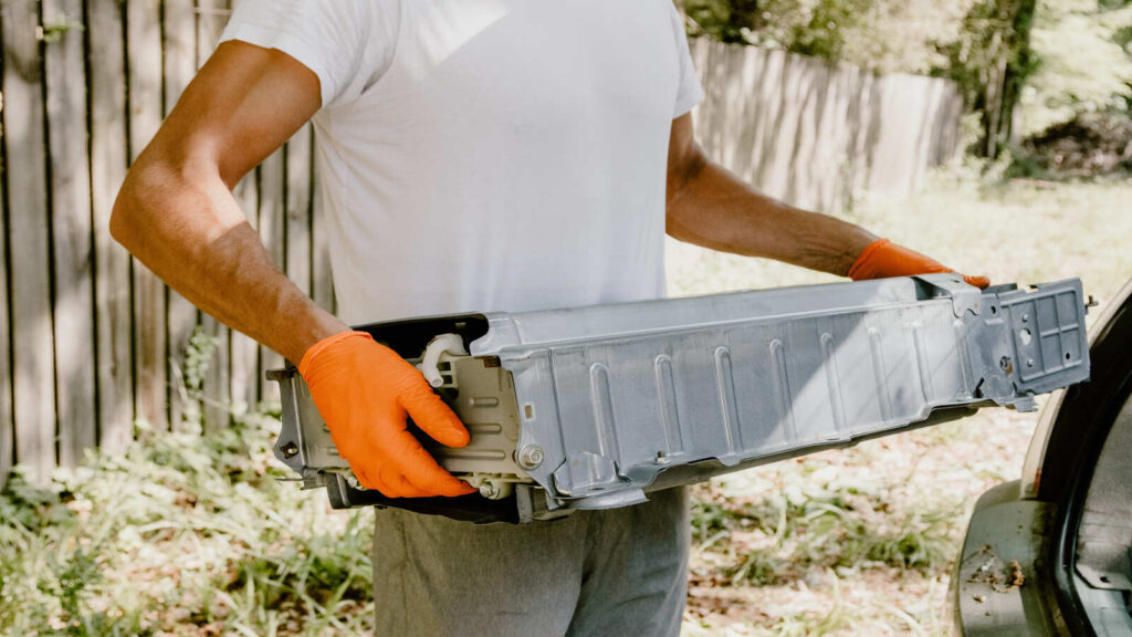 Man holding a Toyota hybrid battery next to a car for maintenance or replacement